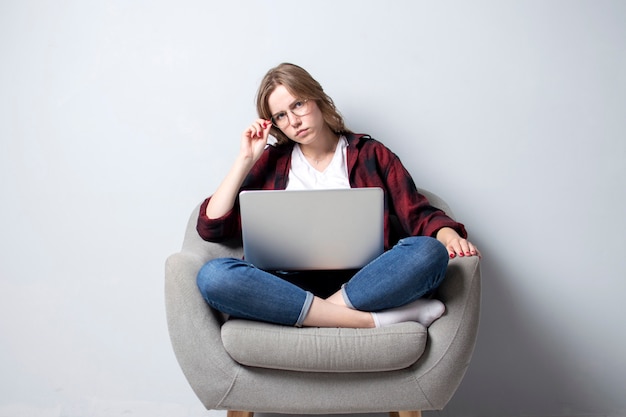 Poważna dziewczyna z laptopem siedzącym na miękkim wygodnym krześle i patrząca w kamerę, kobieta korzystająca z komputera na białej pustej ścianie, freelancing i poprawiająca okulary, miejsce