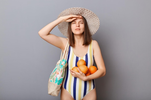 Poważna kobieta ubrana w strój kąpielowy odizolowana na szarym tle, stojąca z pomarańczami, trzyma rękę blisko czoła, patrząc daleko, czekając na swoje tropikalne wakacje