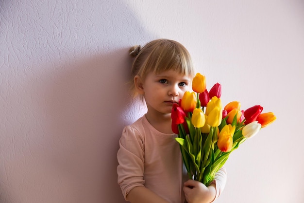 Poważna mała blondwłosa dziewczyna trzyma w dłoniach bukiet kolorowych tulipanów w słonecznym pokoju