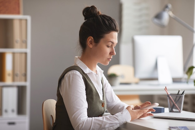 Poważna młoda bizneswoman koncentruje się na swojej pracy online na telefonie komórkowym siedząc w biurze