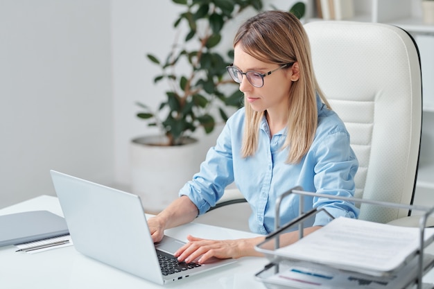 Poważna Młoda Bizneswoman W Niebieskiej Koszuli I Okularach Przegląda Dane Na Wyświetlaczu Laptopa I Dodaje Inne Informacje W Dokumencie