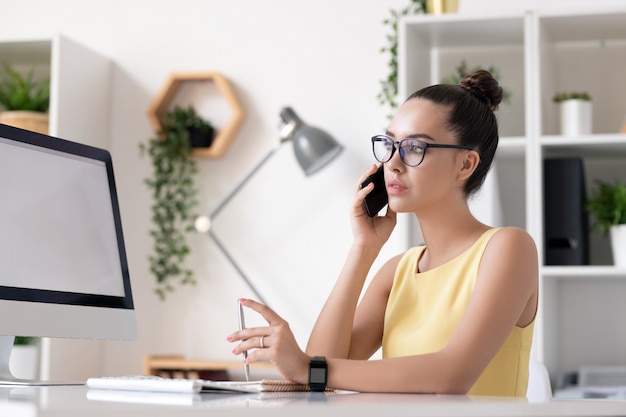 Poważna młoda bizneswoman w okularach siedzi przy biurku i skręcanie pióra podczas rozmowy z klientem przez telefon
