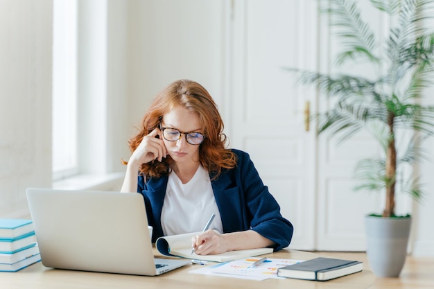 Poważna, pewna siebie przedsiębiorczyni w eleganckim stroju zapisuje informacje, podczas gdy ogląda webinarium biznesowe notuje główne idee siedzi w miejscu pracy przed otwartym komputerem robi notatki dla startupu