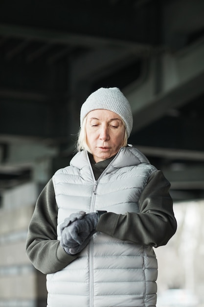 Poważna Starsza Kobieta Rasy Kaukaskiej W Szarym Kapeluszu I Rękawiczkach, Stojąca Pod Mostem I Sprawdzająca Puls Na Trackerze Po Biegu