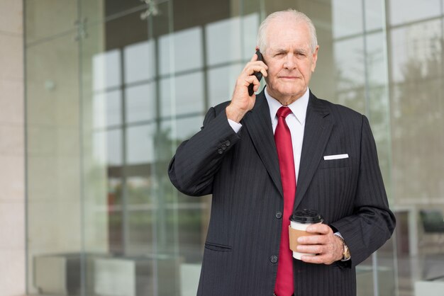 poważne starszy człowiek biznesu wzywając smartphone i trzymając kubek jednorazowy