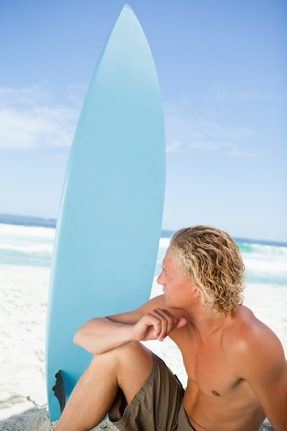 Poważny blondynka mężczyzna obsiadanie na plaży z jego surfboard obok on