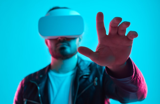 Poważny brodaty mężczyzna w skórzanej kurtce i goglach VR dotyka wirtualnego obiektu podczas interakcji z wyimaginowanym interfejsem w studio z neonami