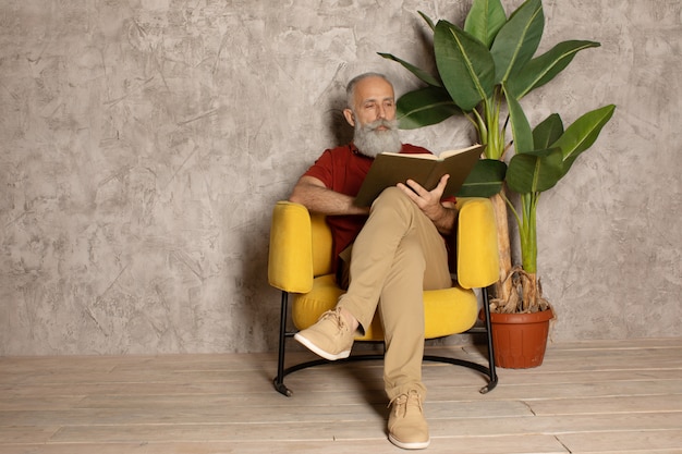 Poważny brodaty starszy mężczyzna czyta ciekawą książkę podczas gdy umieszczający na wygodnej kanapie w mieszkaniu.