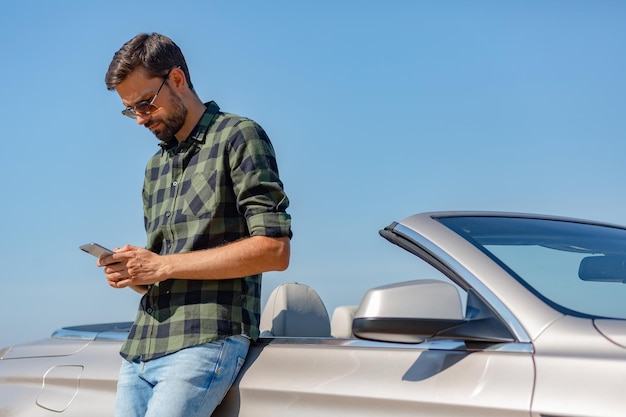 Poważny facet stojący przy samochodzie i patrzący w smartfonie w ciągu dnia