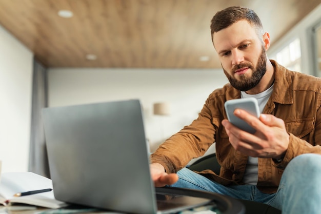 Poważny freelancer używający telefonu i laptopa w domu