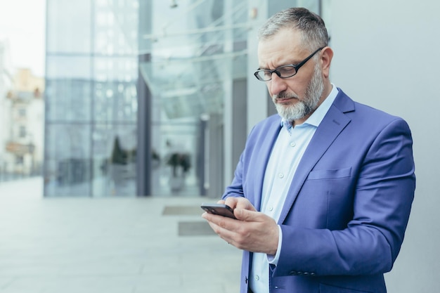 Poważny i zamyślony starszy siwy mężczyzna na zewnątrz budynku biurowego, patrzący na ekran smartfona