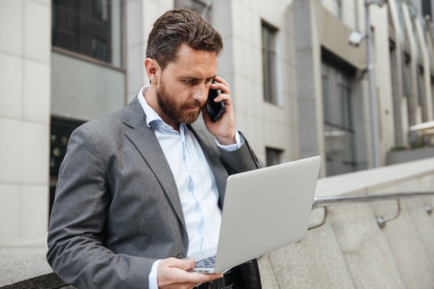 poważny mężczyzna biurowy w klasycznym garniturze pracuje na srebrnym laptopie i rozmawia przez telefon komórkowy, stojąc w pobliżu centrum biznesowego