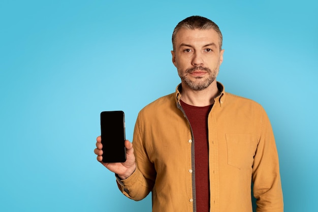 Poważny mężczyzna pokazujący smartfon z pustym ekranem na niebieskim tle