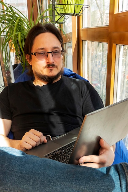 Poważny mężczyzna siedzi na krześle z niebieską fasolą i korzysta z laptopa w domowym biurze na balkonie