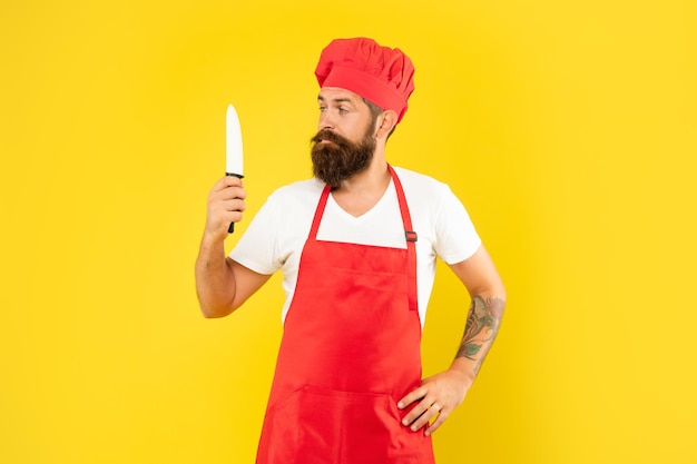 Poważny mężczyzna w czerwonym fartuchu i toczku patrzący na szefa kuchni nóż żółty tło, szef kuchni