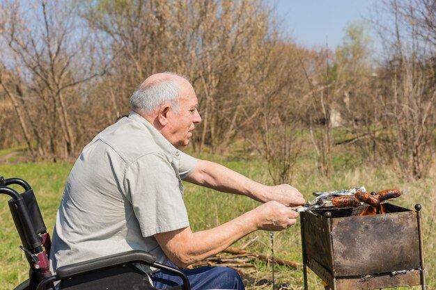 Poważny Mężczyzna W Podeszłym Wieku Siedzi Na Wózku Inwalidzkim, Grillowanie Kiełbasek W Parku W Bardzo Słoneczny Dzień. Zrobione W Widoku Z Boku.