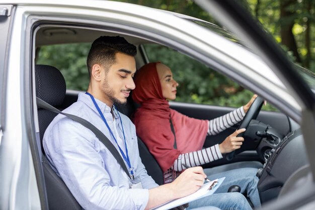 Poważny millenialsi Arab, biorący egzamin na prawo jazdy od kobiety w hidżabie, siedzącej za kierownicą