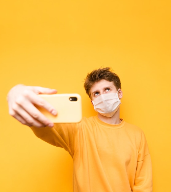 Poważny młody człowiek w masce przeciw koronawirusowi w żółtym swetrze robi selfie na smartfonie