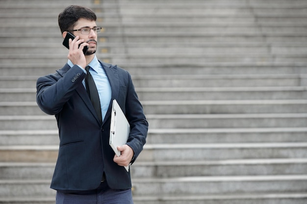 Poważny młody przedsiębiorca stojący na zewnątrz ze schowkiem i rozmawiający przez telefon z kolegą