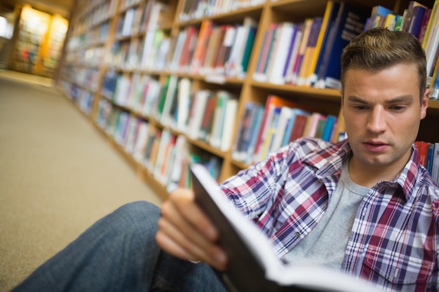 Poważny młody studencki obsiadanie na bibliotecznej podłogowej czytelniczej książce