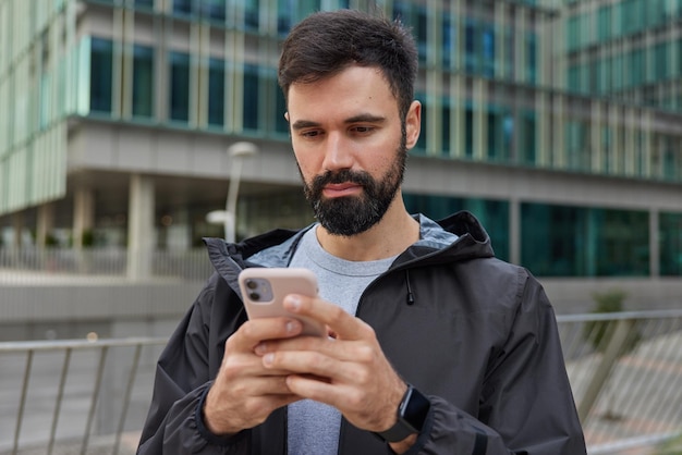 Poważny przystojny facet trzyma telefon komórkowy uważnie patrzy na ekran wysyła wiadomość do znajomych w sieciach społecznościowych pobiera mapy aplikacja pozuje w pobliżu nowoczesnego budynku w mieście nosi smartwatch z anorak