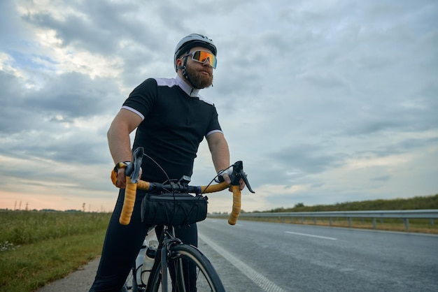 Poważny sportowy mężczyzna z brodą w okularach sportowych i hełmie stojący z rowerem przy drodze