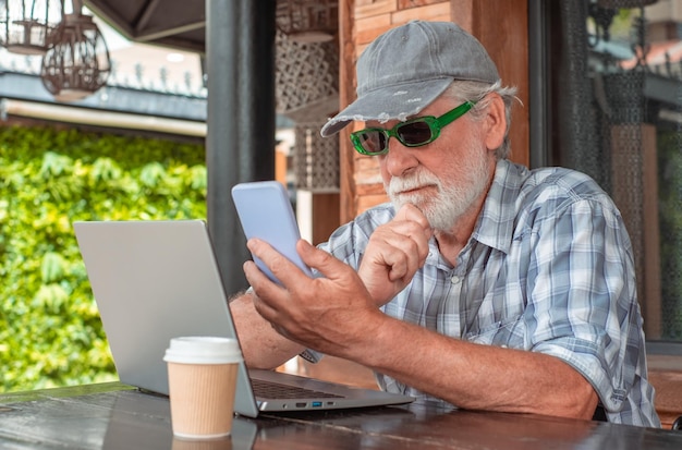 Poważny starszy brodaty mężczyzna w kapeluszu i okularach przeciwsłonecznych używający telefonu do czytania wiadomości siedzący na zewnątrz na drewnianym stole Przystojny starszy mężczyzna pracujący na laptopie