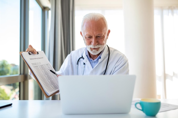 Poważny stary dojrzały profesjonalny lekarz używający laptopa w gabinecie szpitalnym prowadzący szkolenie w zakresie webinarów medycznych pisanie sprawozdania z opieki zdrowotnej konsultowanie się z pacjentem online na spotkaniu telemedycznym