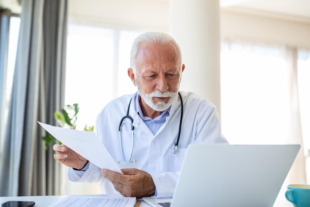 Poważny stary dojrzały profesjonalny męski lekarz korzystający z laptopa w biurze szpitalnym mający medyczne seminarium internetowe pisanie w raporcie opieki zdrowotnej konsultowanie pacjenta online na spotkaniu telemedycznym