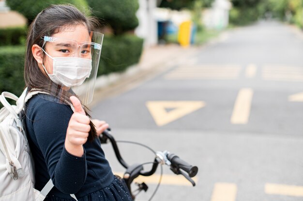 Powrót do szkoły. asian dziecko dziewczyna nosi maskę i daje kciuk z plecakiem na rowerze i idzie do szkoły. pandemia wirusa koronawirusa. Nowy normalny styl życia. Koncepcja edukacji.
