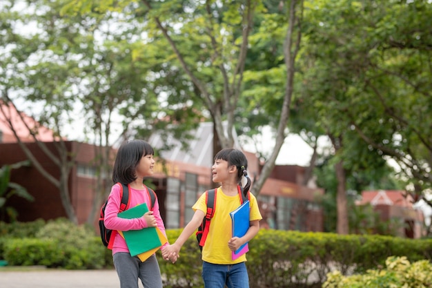 Powrót do szkoły. Dwie śliczne azjatyckie dziewczynki z tornisterem, trzymające książkę i spacerujące razem w szkole
