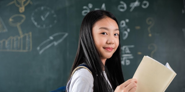 Powrót do szkoły. Portret uśmiechniętej kobiety dziecko ucznia czarnej tablicy kredowej, szczęśliwa piękna azjatycka uczennica stojąca trzymająca książki stojące przed tablicą w klasie, lekcja edukacji