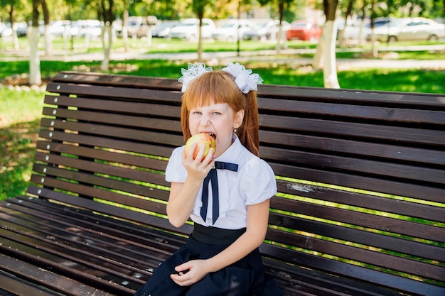 Powrót do szkoły. Śliczna mała uczennica siedzi na ławce na dziedzińcu szkolnym i gryzie zielone jabłko. Właściwe posiłki szkolne na lunch. Mała dziewczynka idzie do pierwszej klasy. Dzień Wiedzy.