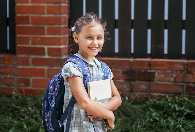 Powrót do szkoły Urocza mała uczennica w sukience z warkoczykami i dużym niebieskim plecakiem trzyma książki i spaceruje po szkolnym dziedzińcu Mała dziewczynka idzie do pierwszej klasy