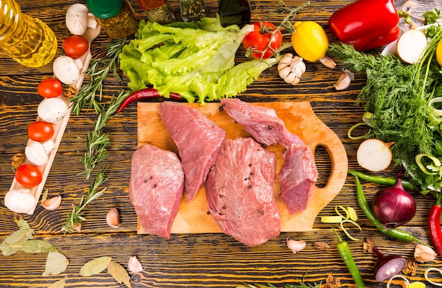 Powyżej widok czterech kawałków surowego czerwonego mięsa na drewnianym stole w otoczeniu cebuli, pomidorów, grzybów i innych warzyw