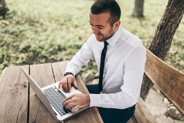 Powyżej widok inteligentnego studenta noszącego białą koszulę, piszącego na komputerze i studiującego Przystojny mężczyzna pracujący na laptopie na drewnianym stole na świeżym powietrzu w parku Technologia zawód i koncepcja edukacji