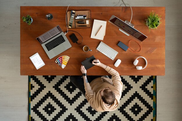 Powyżej widok projektanta interfejsu w bluzie z kapturem siedzącego przy drewnianym biurku pełnym urządzeń i rysującego szkic na tablecie digitizera