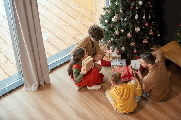 Powyżej widok rodziny z dwójką dzieci siedzących na podłodze w pobliżu choinki i potrząsających pudełkami z prezentami...