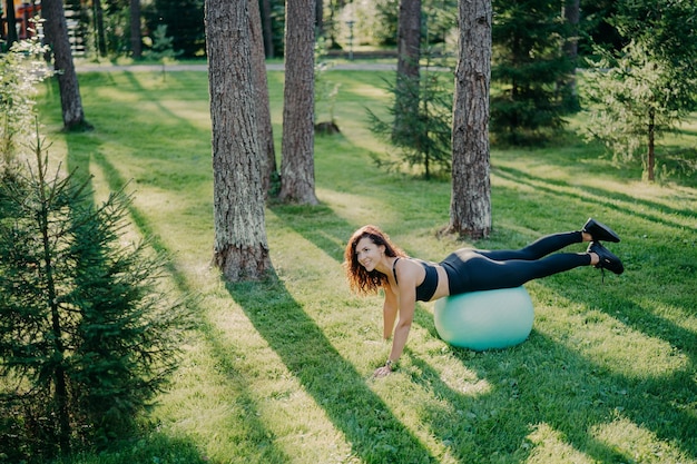 Powyżej widok zdeterminowanej młodej brunetki Europejki ćwiczącej pilates w zielonym parku balansuje na piłce fitness ubranej w odzież sportową wygląda szczęśliwie gdzieś poza nią Selektywna ostrość