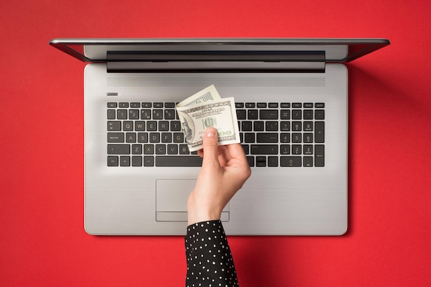 Powyżej zdjęcie szarego laptopa i dłoni trzymającej sto dolarów na białym tle na czerwonym tle