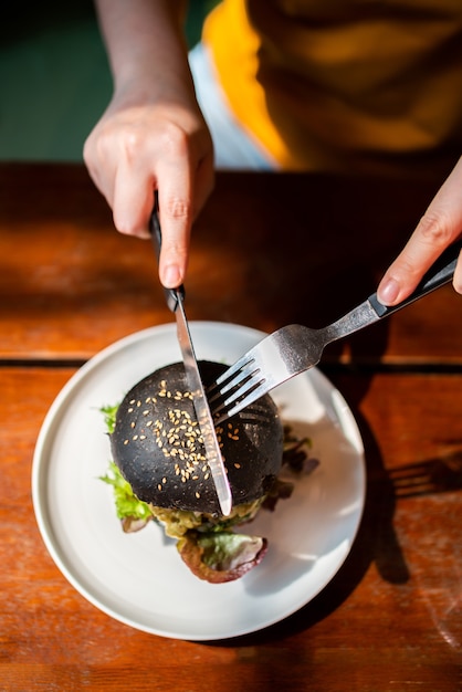 Powyższy Widok Dłoni Kroi Burger Z Brokułami I Komosą Ryżową, Zwieńczony Guacamole, Salsą Z Mango I świeżą Sałatą Nożem I Widelcem, Podawany Na Białym Talerzu. Kreatywny Posiłek Wegański Dla Wegetarian.