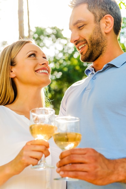 Pozdrawiam kochać! Niski kąt widzenia szczęśliwej młodej pary kochającej trzymającej szklanki z białym winem i patrzącej na siebie z uśmiechem, stojąc razem na zewnątrz