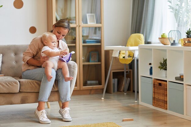 Poziome strzał nowoczesnej kobiety rasy kaukaskiej sobie dorywczo strój siedzi na kanapie z dzieckiem na kolanach, grając z zabawkami