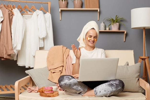 Poziome ujęcie atrakcyjnej kaukaskiej kobiety ubranej w białe szare spodnie z koszulką i ręcznikiem na głowie, siedzącej na kaszlu i używającej laptopa do rozmowy wideo machającej ręką i witającej się