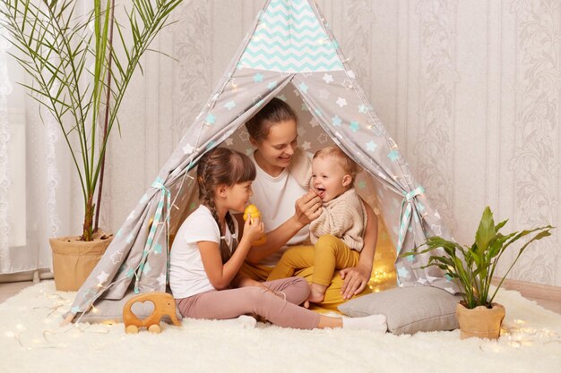 Poziome ujęcie atrakcyjnej kobiety siedzącej na podłodze w namiocie tipi z jej uroczymi dziećmi ludzi wyrażających pozytywne emocje, śmiejąc się, ciesząc się spędzaniem czasu razem