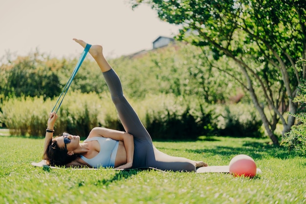 Poziome ujęcie elastycznej kobiety podnosi nogę i wykonuje ćwiczenia z gumą fitness leżącą na karemacie, nosi legginsy, a przycięty top ma idealną figurę pozuje na zewnątrz na zielonej trawie Sportsmenka używa ekspandera