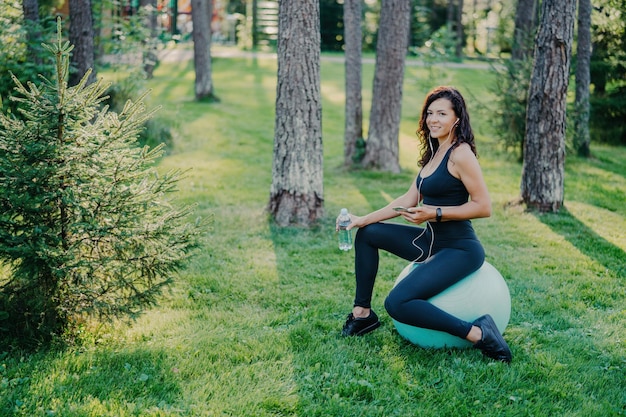 Poziome ujęcie europejskiej brunetki ubranej w przycięty top i legginsy siedzi na piłce fitness używa smartfona i słucha muzyki w słuchawkach trzyma butelkę świeżej wody pozuje w lesie