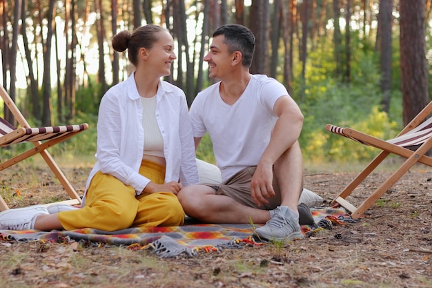 Poziome ujęcie kochającej się pary siedzącej w parku na ziemi i patrzącej na siebie z miłością, wyrażającą romantyczne uczucia, noszące zwykłe ubrania, odpoczywające w lesie