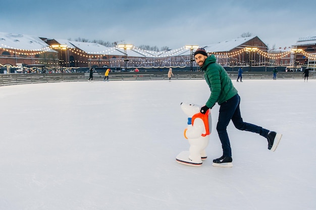 Poziome ujęcie mężczyzny nosi ciepłą czapkę gree anorak i łyżwy uczy się jeździć na łyżwach na lodzie korzysta z pomocy łyżwiarskiej cieszy się wolnym czasem i feriami zimowymi uśmiecha się szczęśliwie do kamery Koncepcja hobby i ludzi