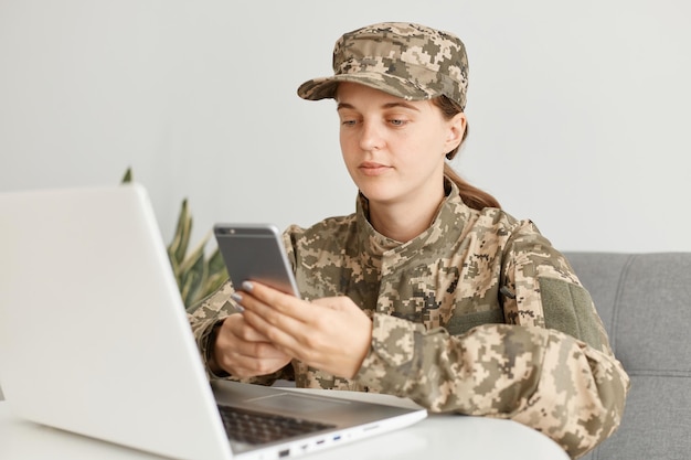Poziome ujęcie spokojnej kobiety żołnierza w mundurze kamuflażu i kapeluszu, pozowanie w domu, siedząc przed komputerem przenośnym i przy użyciu telefonu komórkowego, ma spokojną ekspresję.